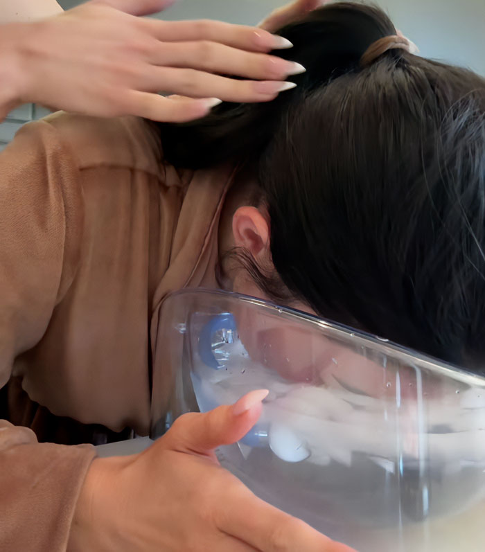 Person with dark hair in a brown robe using an ice water bowl as part of a Botox beauty hack routine at home.