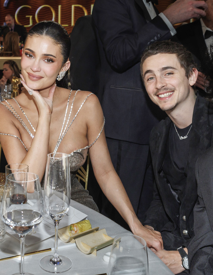 Kylie Jenner at Golden Globes seated at table with drinks and holding hands with a smiling man during event.