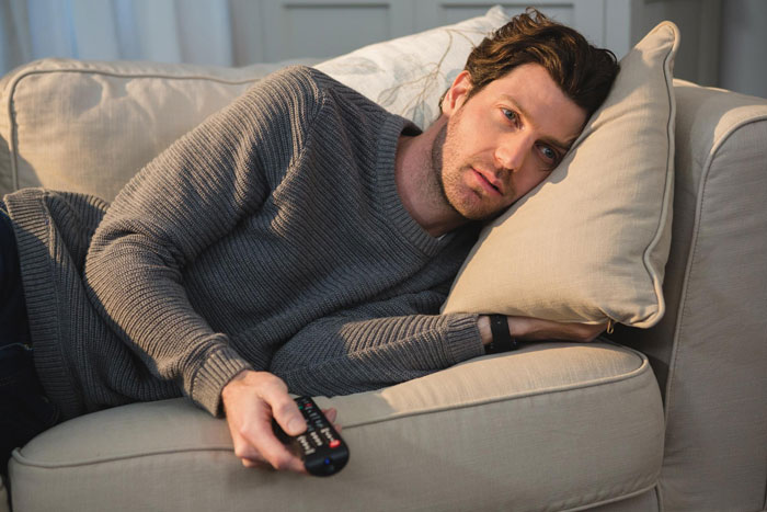 Man in gray sweater lying on couch holding remote control, illustrating fed up wife and useless hubby energy conflict.