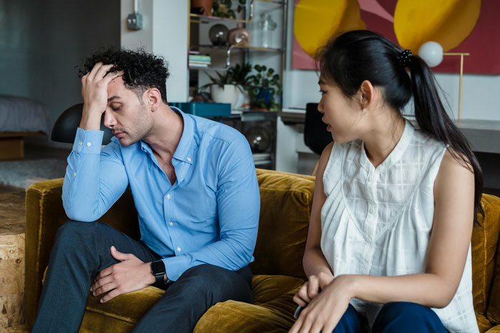 Couple having tense conversation on couch, wife matching useless hubby&rsquo;s energy, both showing frustration and distress.