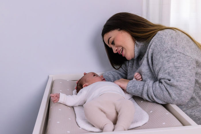 Woman in gray sweater smiling at baby lying on changing table, illustrating man offers to raise pregnant ex baby story.