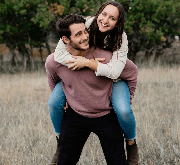 Young couple outdoors with the woman riding piggyback on the lying boyfriend in a casual field setting.