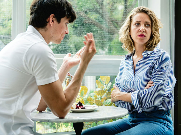 Young woman showing distrust as lying boyfriend explains a trip while sitting at a cafe table near a window.