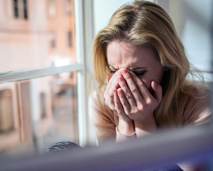 Woman upset by lying boyfriend who went on a trip with a female friend, sitting by the window covering her face.