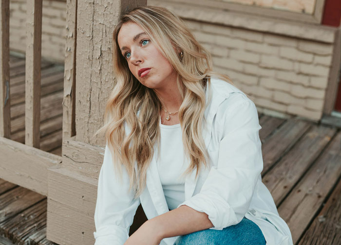 Young woman looking thoughtful and upset, sitting on wooden porch, relating to lying boyfriend who went on trip with female friend