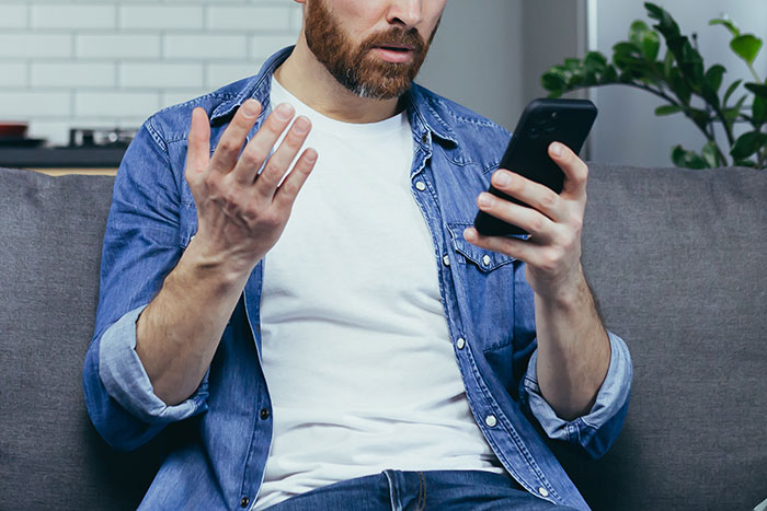 Man sitting on couch looking shocked and confused while reading a message on his smartphone about wife&rsquo;s cheating.
