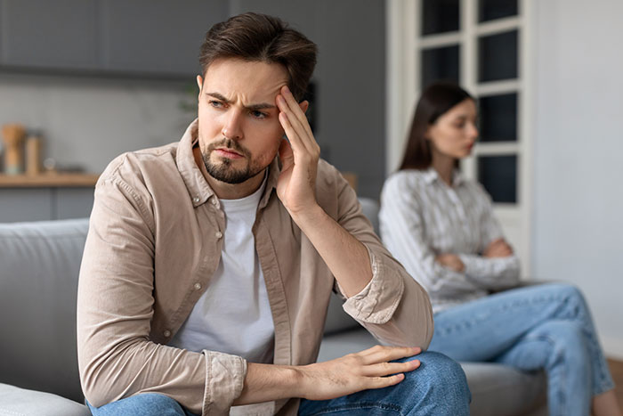 Man sitting on couch looking distressed while wife sits upset and distant in the background after cheating revelation.