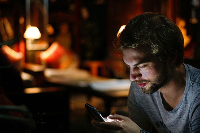 Young man smiling while reading lovey-dovey texts on his phone during a dimly lit evening at home.