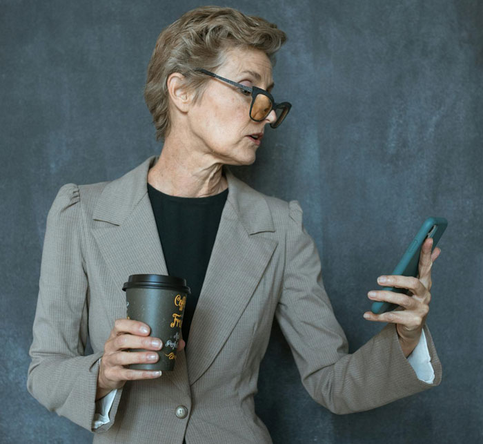 Older woman in glasses holding coffee cup and looking at her phone, portraying suspicion and concern discreetly.
