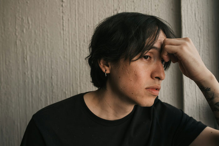 Young man looking disturbed and thoughtful, resting his head on his hand against a textured wall background.