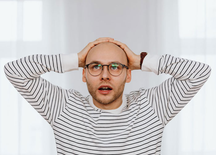 Stressed bald man wearing glasses and striped shirt, holding head in disbelief, representing unemployed and jobless struggles.