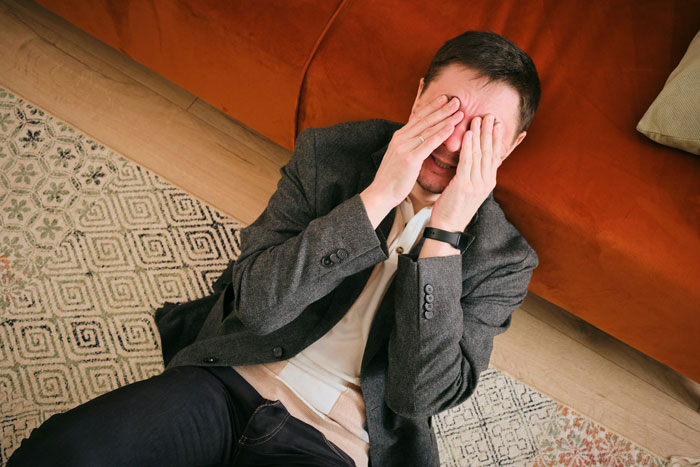 Man wearing a gray blazer sits on the floor, covering his face, reflecting stress about in-laws refusing to help with childcare.