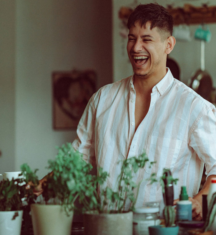 Man laughing at home near plants, illustrating in-laws refusing childcare help and wanting to move in conflict situation.