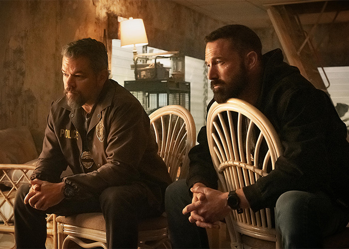 Two men sitting on wicker chairs in a dimly lit room, with serious expressions and casual dark clothing.