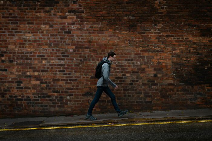 Person walking along a sidewalk with a backpack against a brick wall, illustrating everyday life cheat codes.