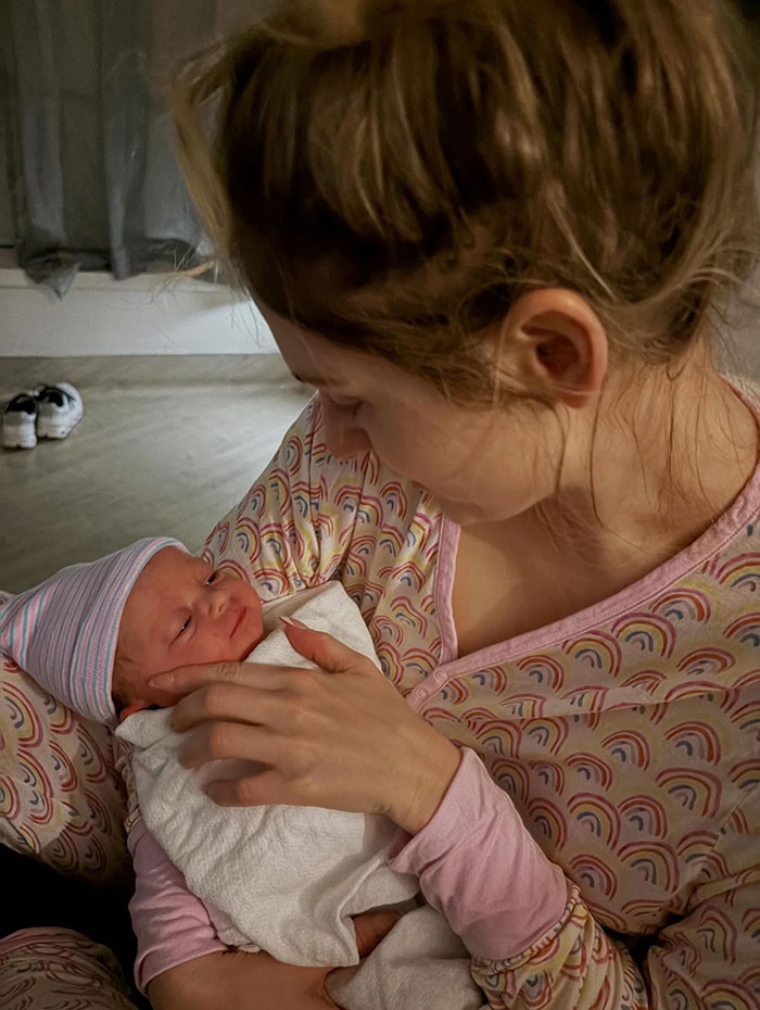 Meghan Trainor cradling newborn baby wrapped in blanket while wearing rainbow-patterned pajamas in soft indoor lighting.