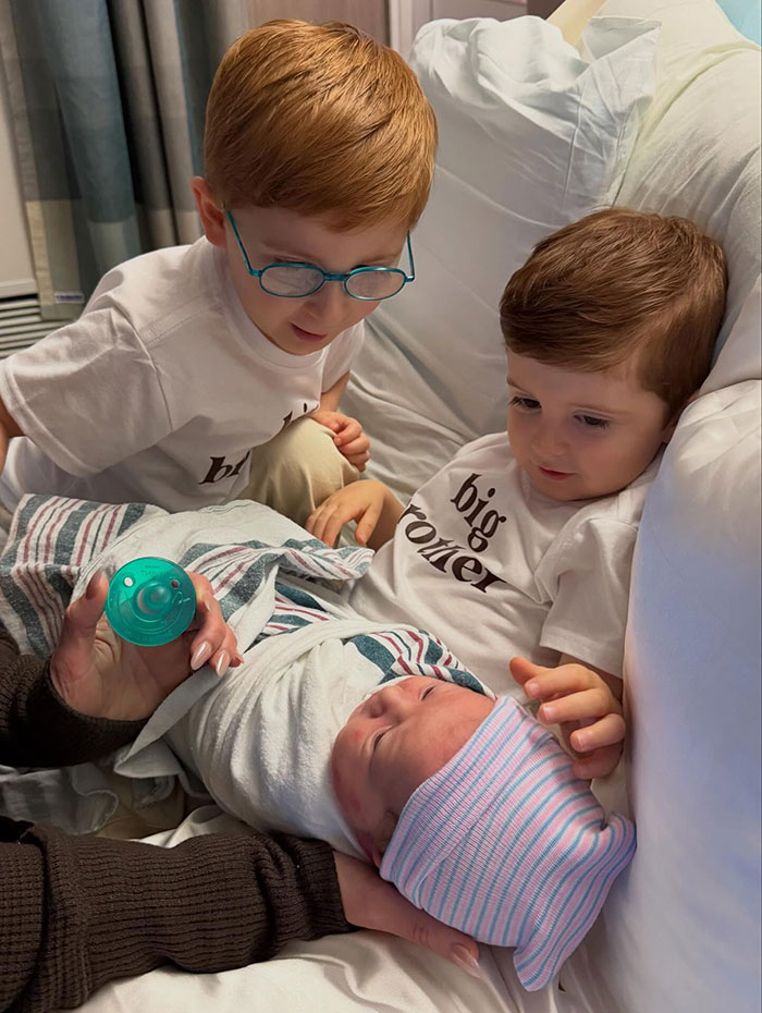 Two young boys wearing big brother shirts look at a newborn baby in a hospital bed with a secret surrogate baby theme.