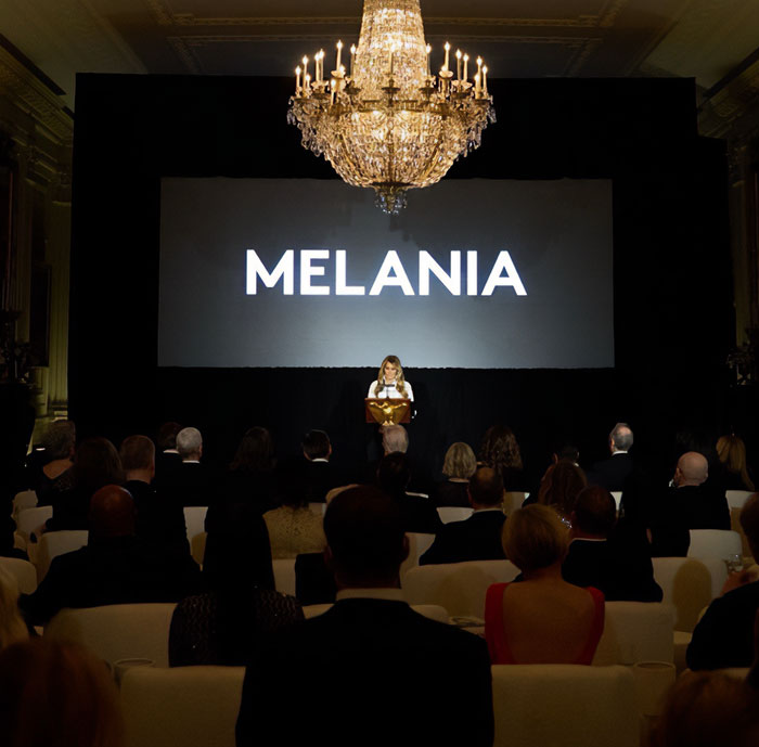Audience watching a presentation about Melania Trump at an event with a large chandelier and dim lighting.
