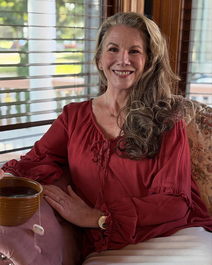 Smiling woman in a red blouse sitting indoors with a cup of tea by the window, relaxed and comfortable at home.