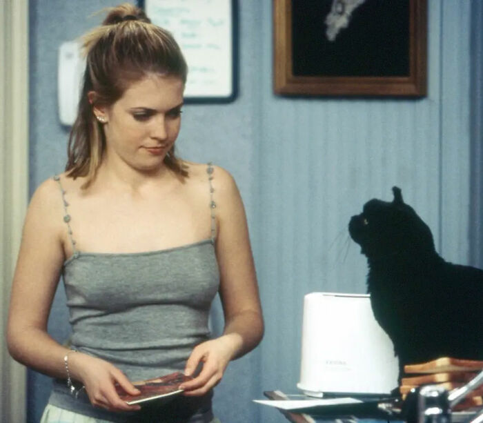 Nickelodeon star in a gray top holding a book near a black cat, reflecting on a humiliating moment with Britney Spears.