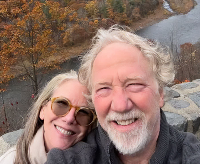 Melissa Gilbert and husband Timothy Busfield smiling outdoors near a river, autumn trees in the background.