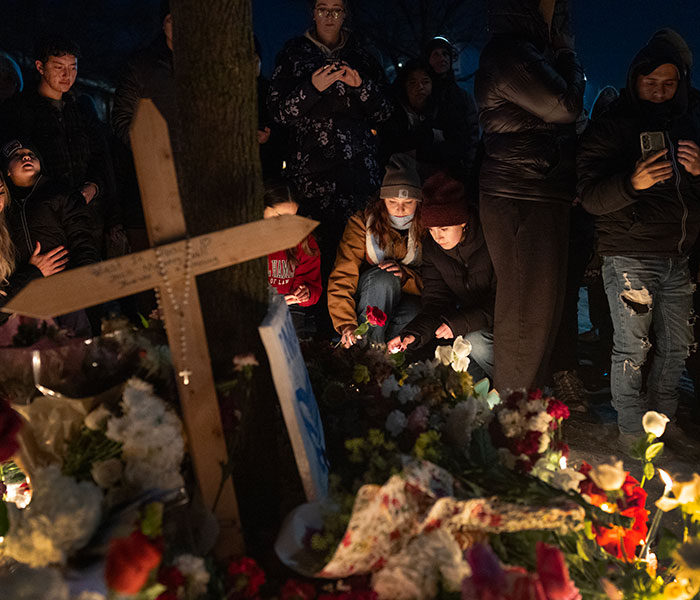 Vigil with people gathered around flowers and a wooden cross, mourning woman slain by ICE in Minneapolis.