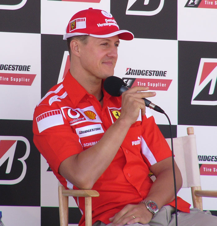 Michael Schumacher in red Ferrari racing gear speaking into microphone at a Bridgestone F1 event press conference.
