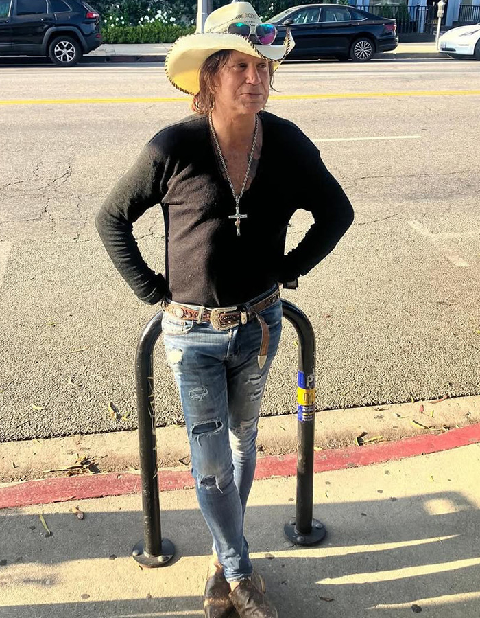 Mickey Rourke wearing a cowboy hat and cross necklace, standing on a sidewalk in casual clothes facing eviction over unpaid rent.