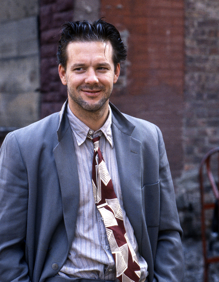 Mickey Rourke dressed in a worn suit and patterned tie, smiling slightly outdoors near an urban brick wall.