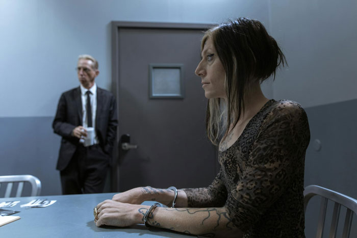 Woman in handcuffs sitting in interrogation room while police officer stands in background during family drama arrest. Woman in handcuffs sitting in interrogation room while police officer stands in background during family drama arrest.