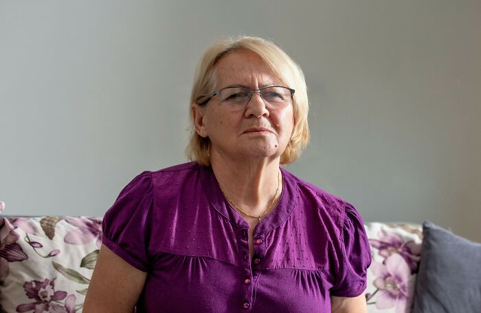 Older woman wearing glasses and purple blouse sitting on floral couch, looking stern and contemplative indoors.