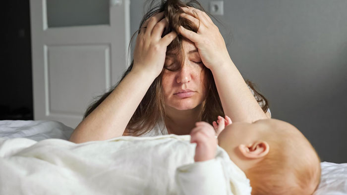 Tired mother resting head in hands while looking at newborn baby lying on bed in a quiet room.