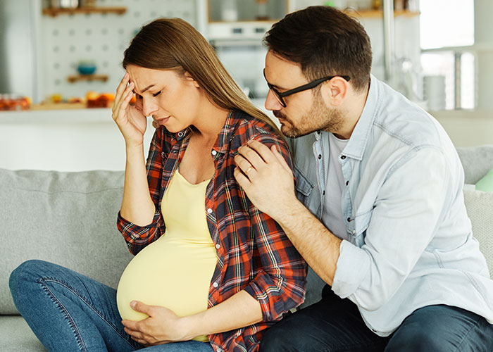 Pregnant woman upset on couch while partner comforts her, highlighting expired food in her pantry from 1999 issue.