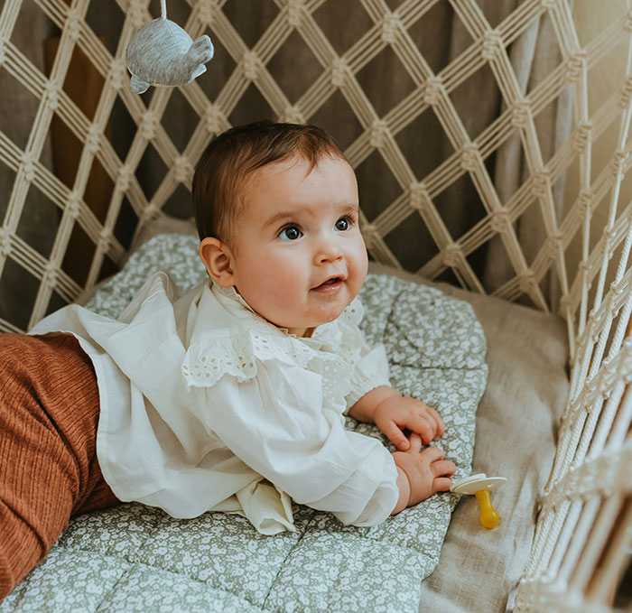Mom takes her baby; infant lying in woven crib looking up, wearing white blouse with pacifier beside her