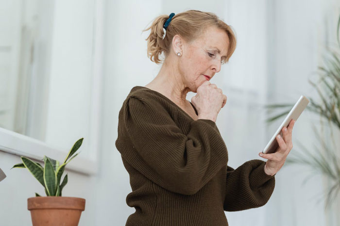 Mature woman thoughtfully using a tablet indoors near potted plant, reflecting on her celebration and independence from controlling vegan friend.