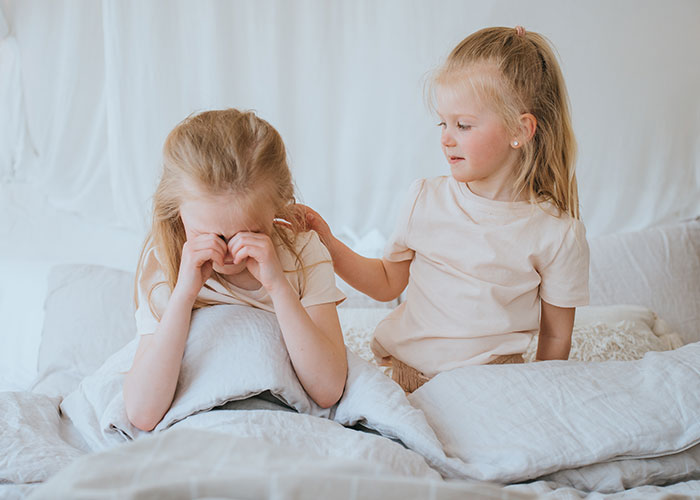 Two young girls in light clothes on a bed, one comforting the other, illustrating mil tells kids parents passed away.