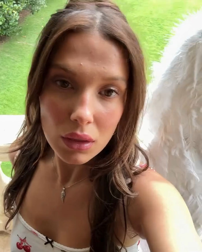 Millie Bobby Brown close-up selfie outdoors wearing angel wings and a cherry print top with natural makeup and loose hair.