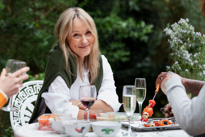 Woman smiling at outdoor dinner with wine and grilled skewers, highlighting irate sister taking mom out alone.