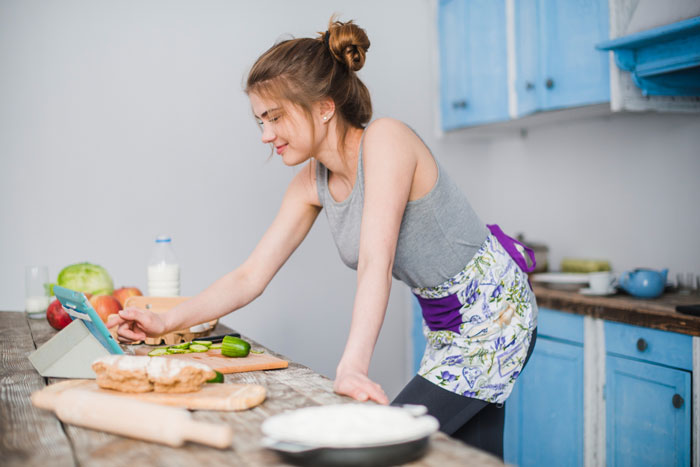 Young woman in apron cooking in kitchen, using a tablet with mom-boyfriend-food-drama theme in focus