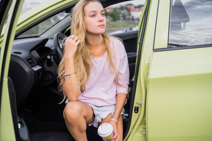 Young woman burned out managing chores and school sitting in car holding coffee, looking stressed and exhausted.