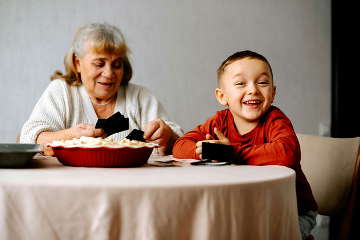 Selfish lady with adopted family member, elderly woman sorting cards while a cheerful boy laughs at a kitchen table