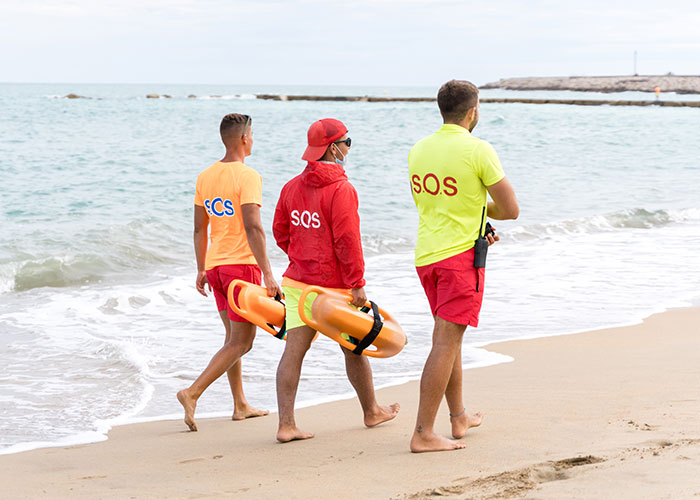 Three lifeguards walking along the beach carrying rescue equipment, highlighting ironic ways people passed away near water.