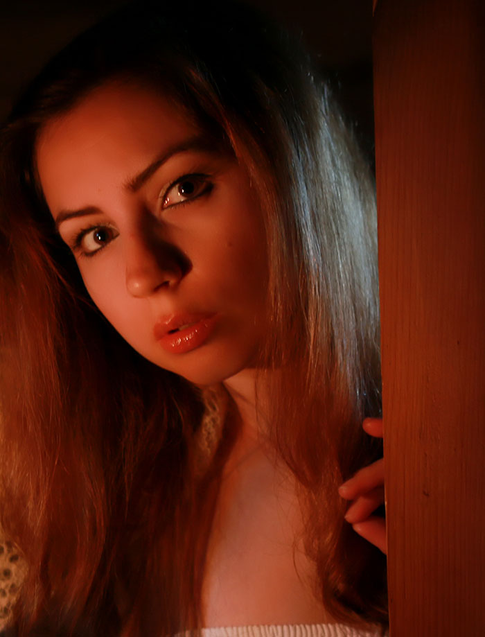 Young woman with an overwhelming feeling, peering nervously from behind a door in a dimly lit setting.