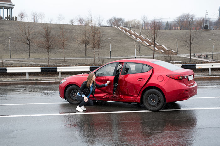 Woman inspecting damaged red car on rainy road, showing overwhelming feeling to leave after an accident situation.