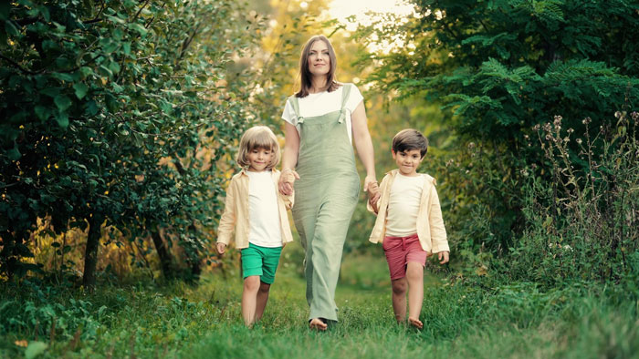 Woman walking with two children in a yard surrounded by trees, illustrating a child-hating hermit neighbor conflict.