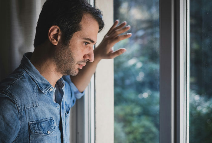 Man in a denim shirt looking out window concerned about nightmare neighbors secretly using family&rsquo;s backyard without permission