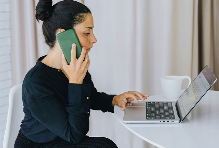 Woman speaking on phone while using laptop, managing complaints about neighbors and family&rsquo;s backyard issues.