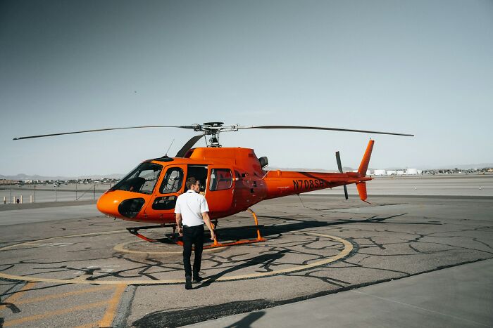 A man walking toward a bright orange helicopter on an airport runway showing how far from reality rich people can be.