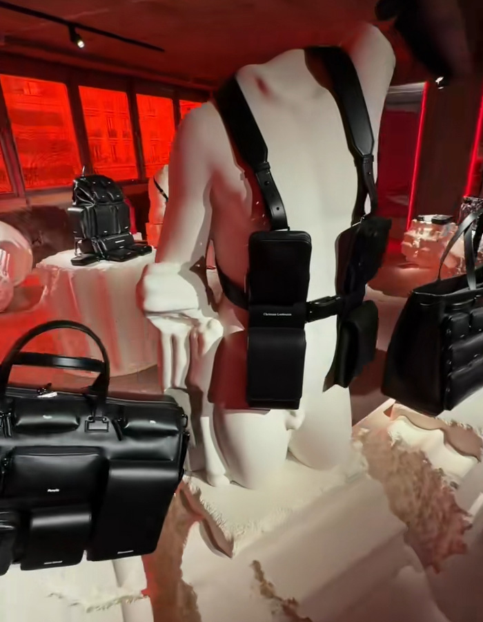 Christian Louboutin black leather bag collection displayed on white mannequins in a red-lit showroom setting. Christian Louboutin black leather bag collection displayed on white mannequins in a red-lit showroom setting.