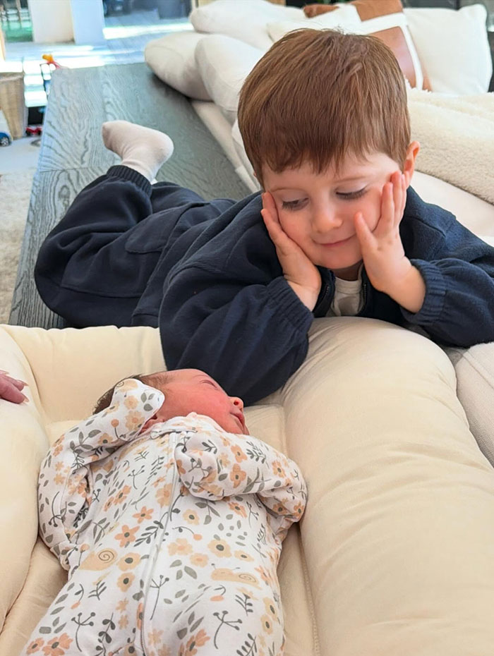 Young child lying next to newborn baby in floral outfit, capturing Meghan Trainor's secret surrogate baby moment performative scene.